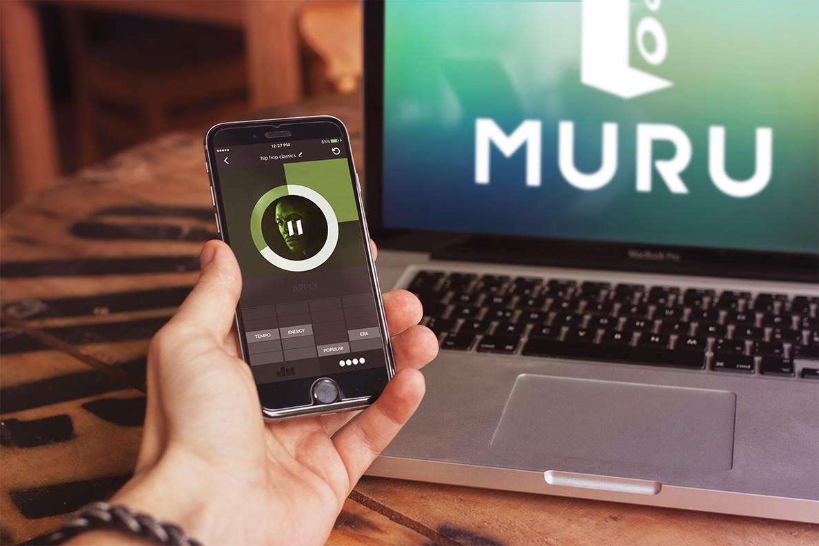 A hand holding a phone showing a playlist called hip hop classics, with a play control and sliders for tempo, energy, popularity and era, in front of a laptop displaying the MURU logo.